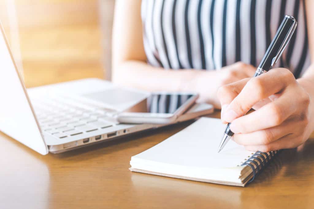Vrouw schrijft met pen op een kladblok bij haar laptop en telefoon